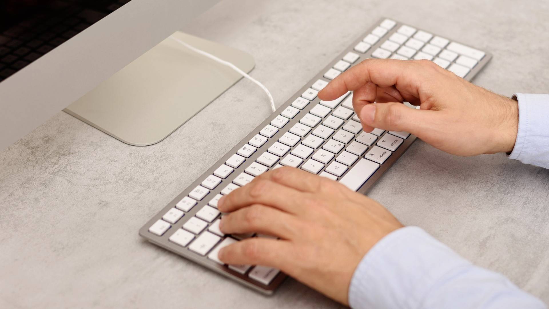 Person, der skriver på et hvidt tastatur foran en computerskærm. Hænderne er fokuseret på indtastning i en arbejdspladsomgivelse.