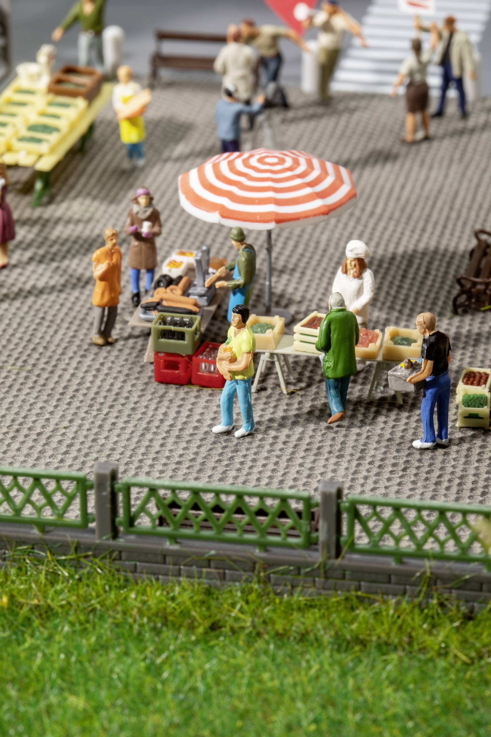 Miniaturudgave af en travl markedsplads med flere figurer ved boder under en rød-hvid parasol.