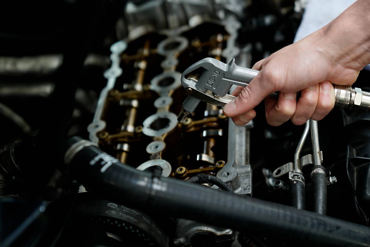 En hånd, der holder en skruenøgle og arbejder på en bildel, formentlig motoren, hvor metalliske komponenter er synlige.