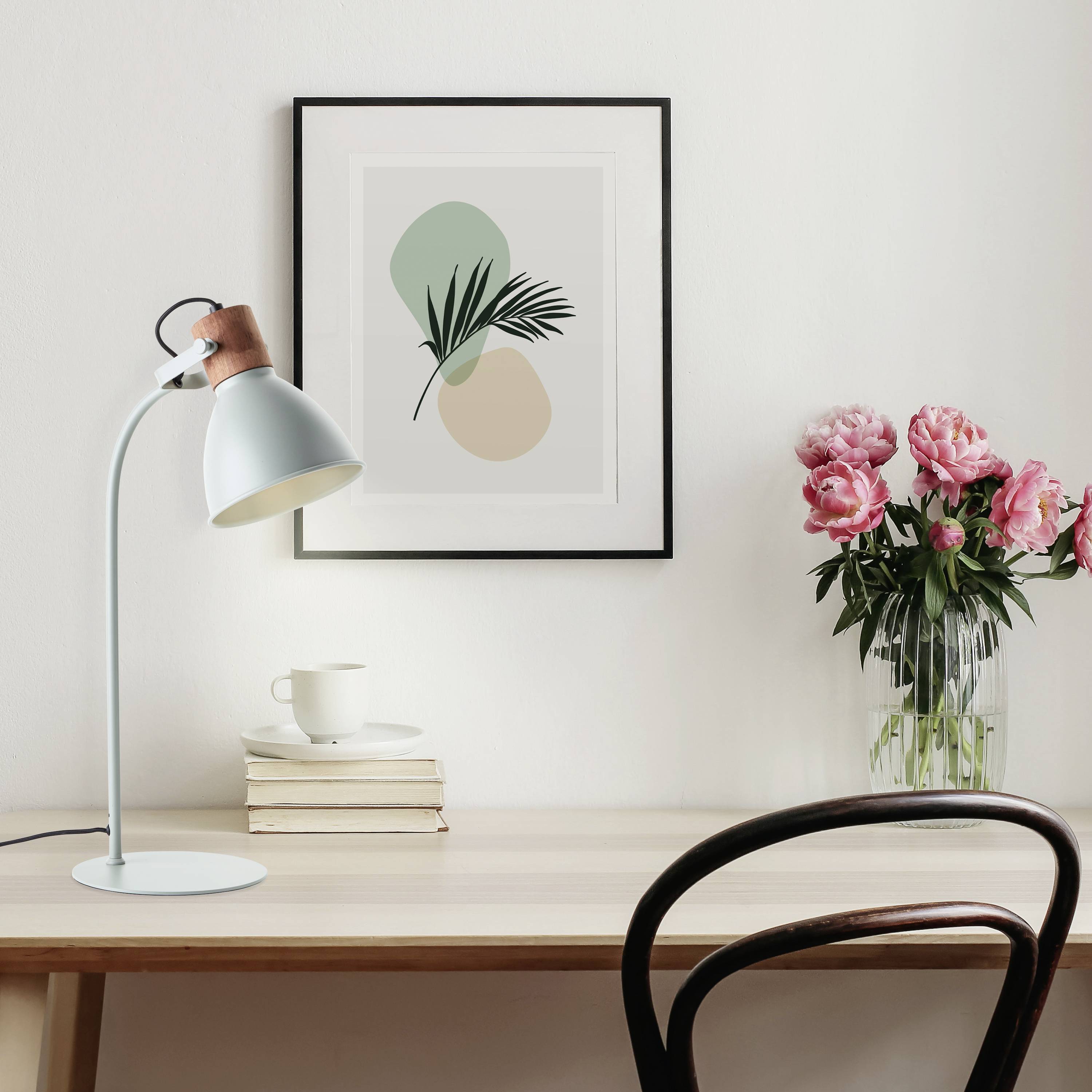 Et skrivebord med en lampe, en bunke bøger og en kop. På væggen hænger et minimalistisk billede. Til højre står en vase med lyserøde blomster.