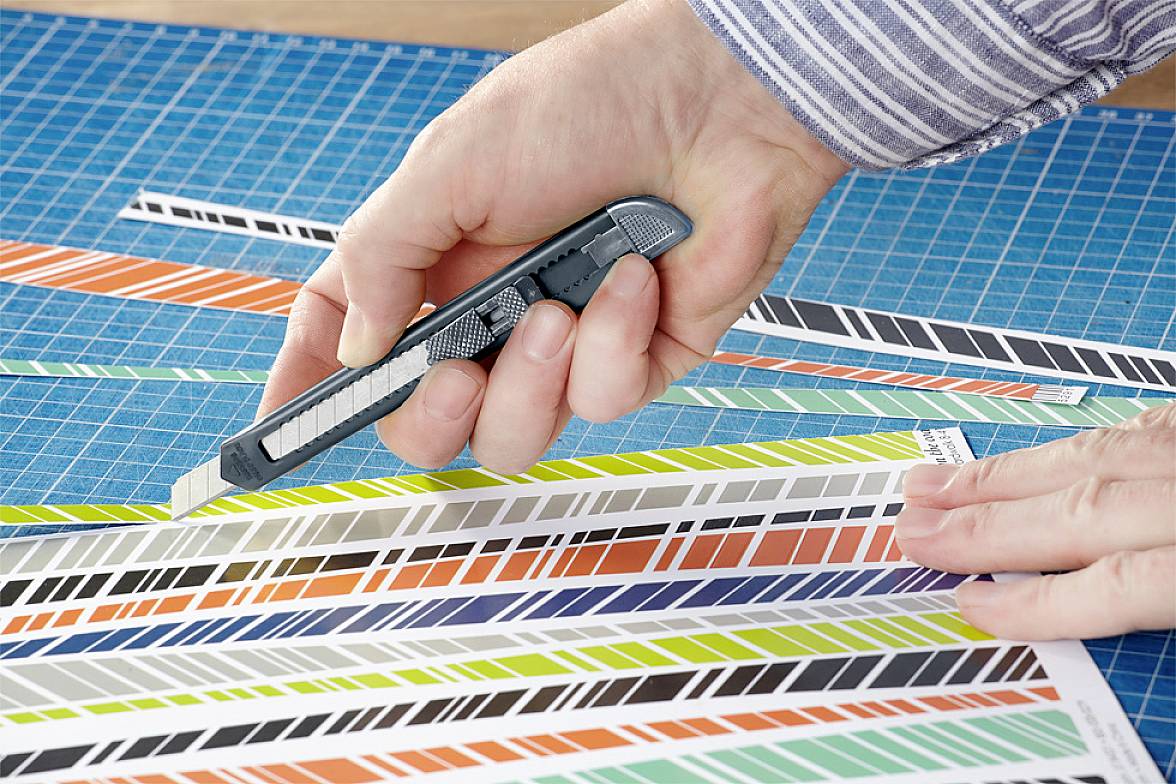 En hånd holder en skærekniv og skærer farvede strimler på en skæremåtte med gitterlinjer.