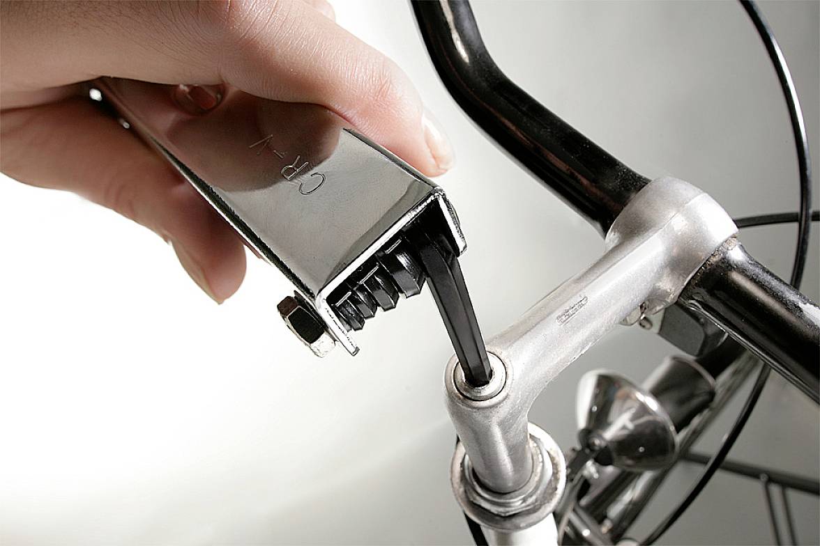 En hånd spænder en skrue på cykelstyret ved hjælp af et nøgleformet værktøj. Styret er delvist synligt.