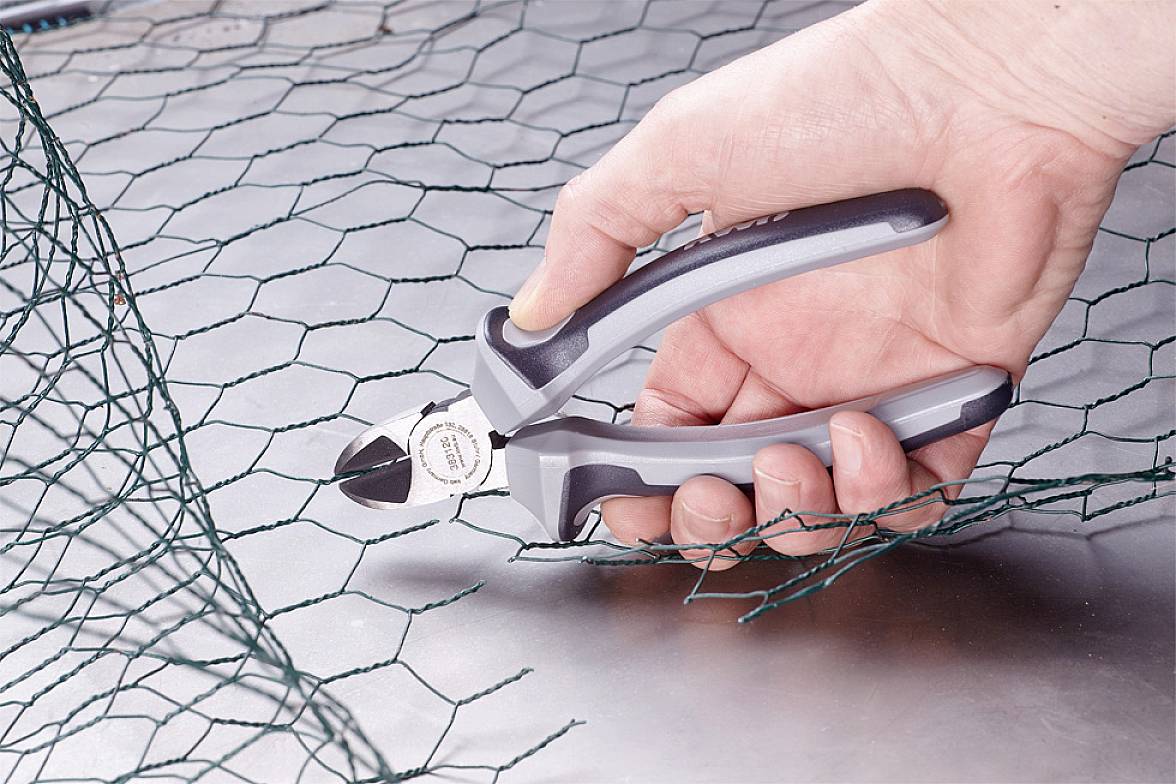 En hånd holder en sidevejsskærer, der klipper grøn trådnet over. Nettet er udlagt på et metalbord.