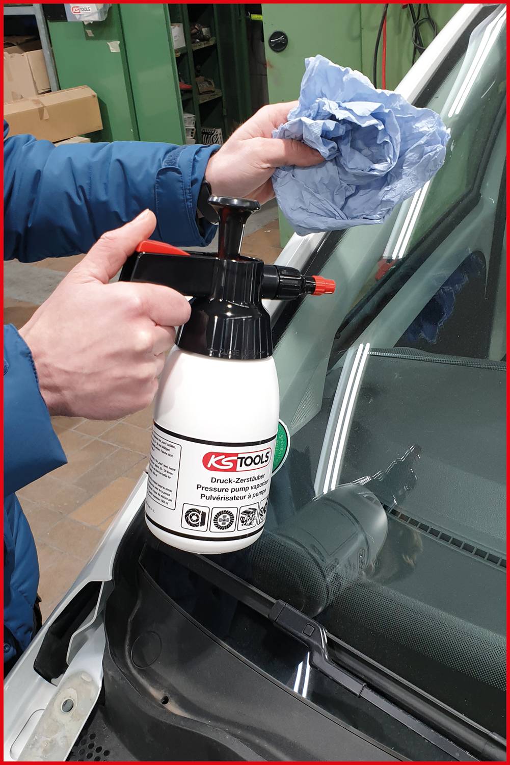 En person sprayer rengøringsmiddel på en forrude med en hvid sprayflaske og tørrer den af med en blå klud.