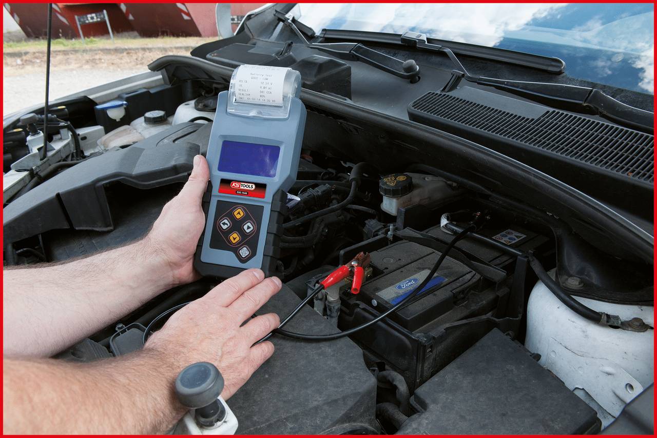 En person bruger en elektronisk diagnoseenhed til at kontrollere bilbatteriet under motorhjelmen. Et batteritestapparat med kabler er tilsluttet.