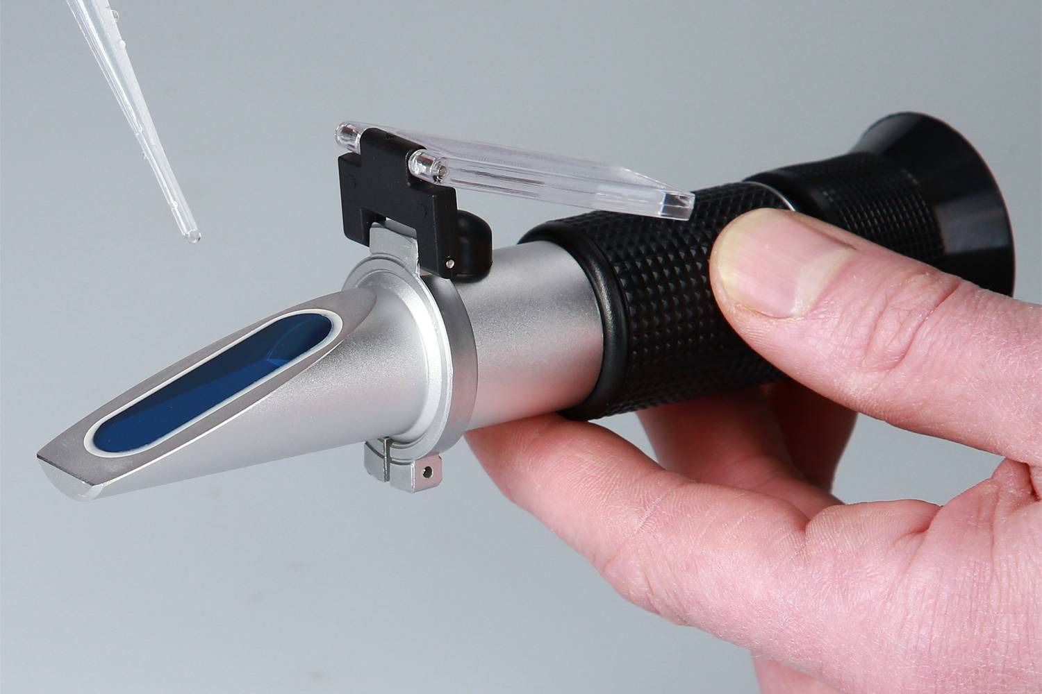 En hånd holder et refraktometer. En pipette drypper væske på synsfeltets overflade for at måle væskens egenskaber.