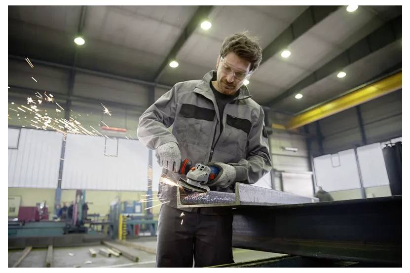 En fabriksarbejder i sikkerhedsudstyr bruger et elektrisk værktøj til at slibe et stykke metal i en industriel værksted, med gnister, der flyver rundt.
