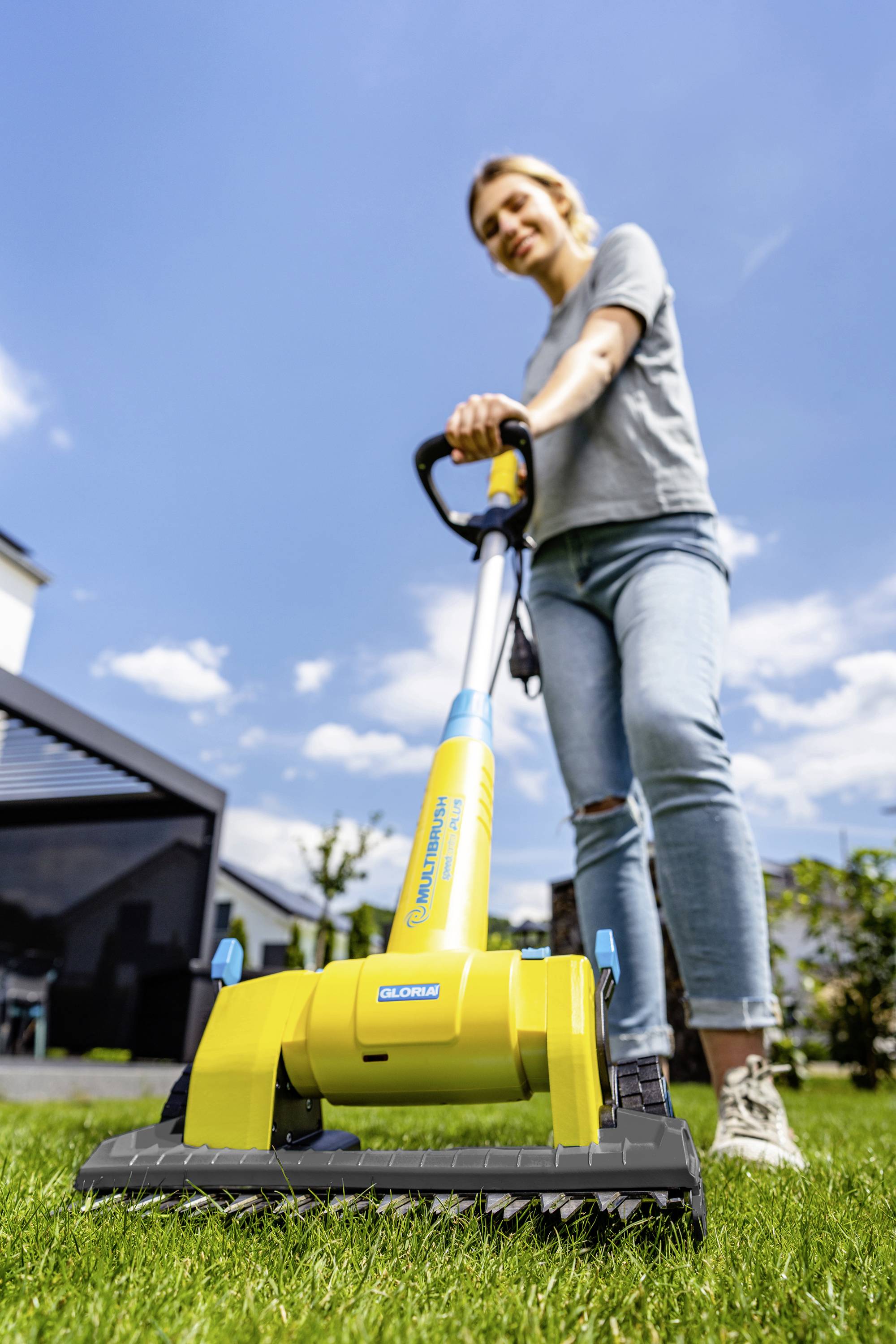 En person slår græsset med en gul elektrisk plæneklipper på en solrig dag foran et moderne hus.