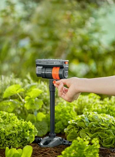 En hånd justerer et sort og orange vandudløb i en have, omgivet af grønne planter og jord, hvilket antyder et vandes-anlæg.