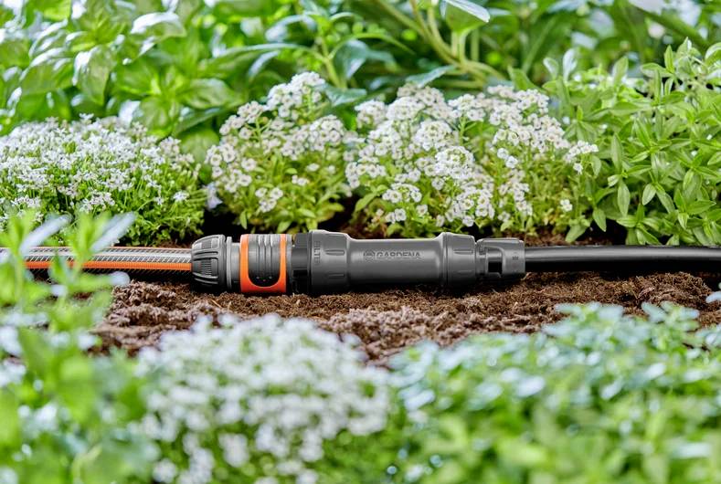 En havevandingssamler er sat fast på en slange, omgivet af frodig grøn vegetation og små hvide blomster, hvilket indikerer effektiv vanding.