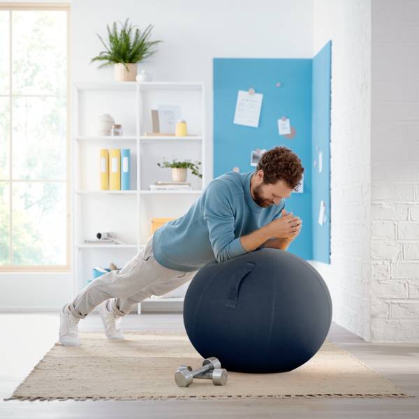 En mand i sportsligt tøj støtter sig op ad en fitnessboldt i stuen. I baggrunden ses hylder og en opslagstavle.
