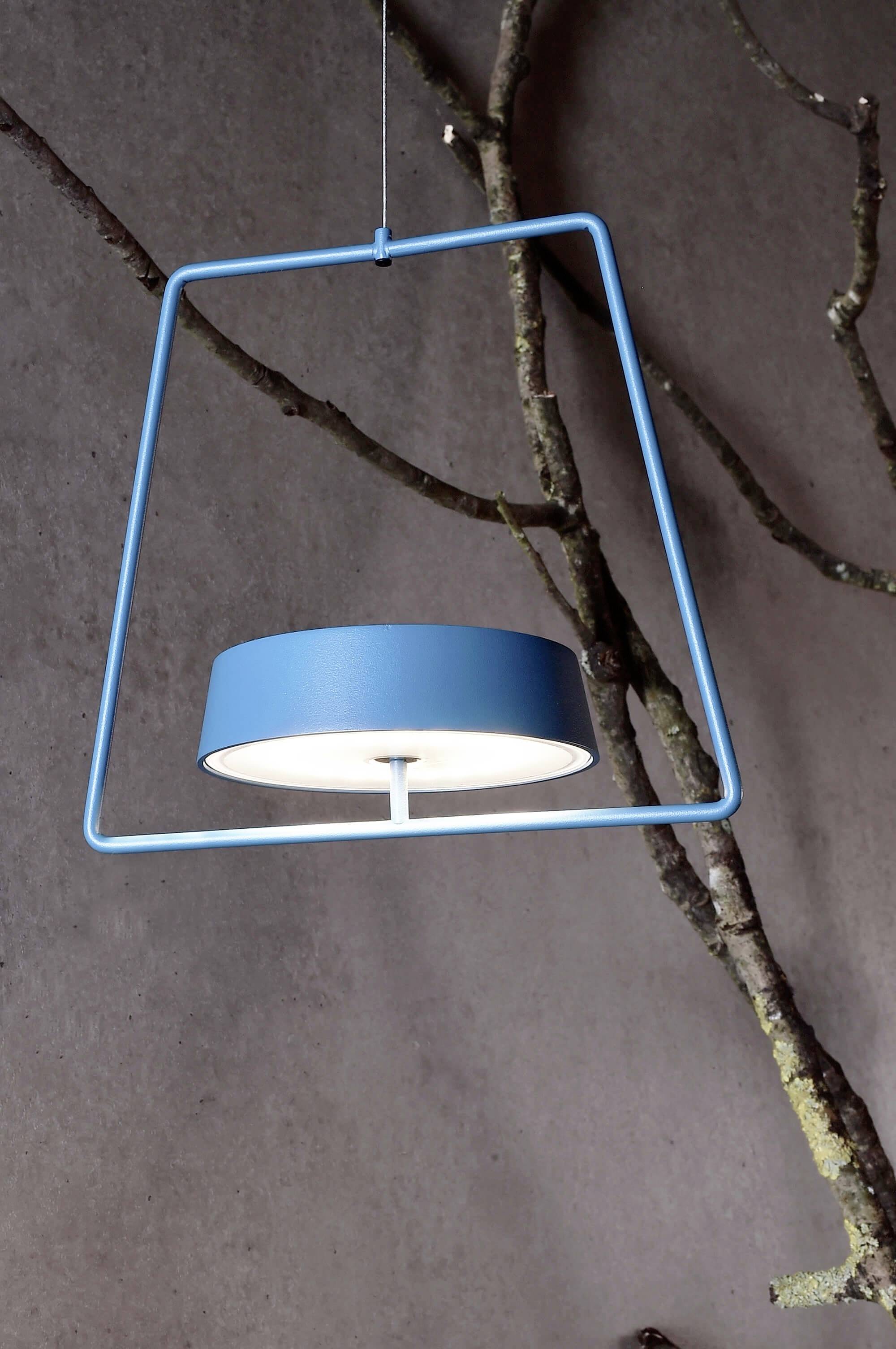 Et blåt, moderne designet pendel, indsat i en minimalistisk, geometrisk ramme, hænger foran en grå væg med grene i baggrunden.
