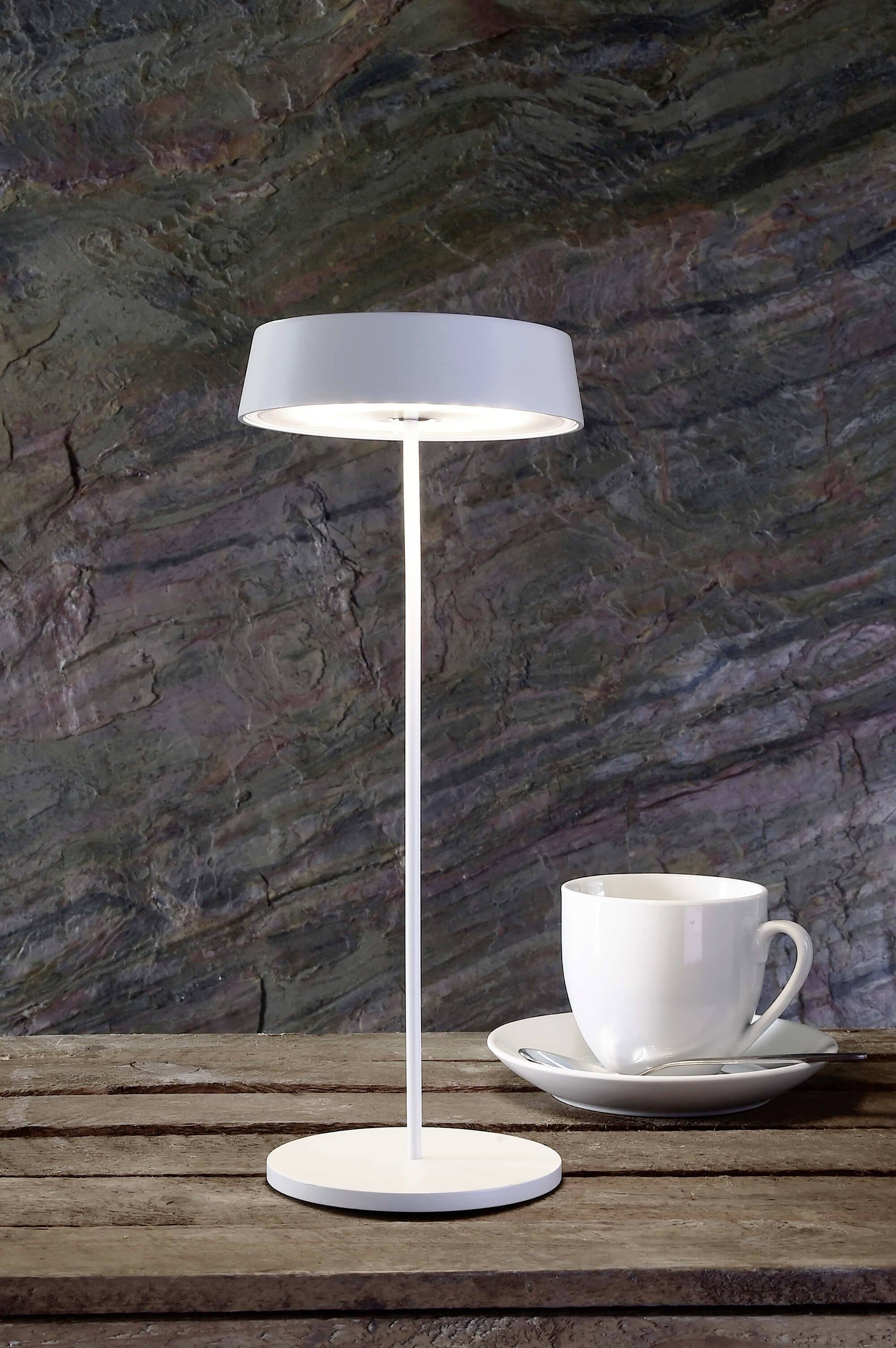 Et hvidt bordlampe og en hvid kop med underkop står på et træbord foran en struktureret stenmur.