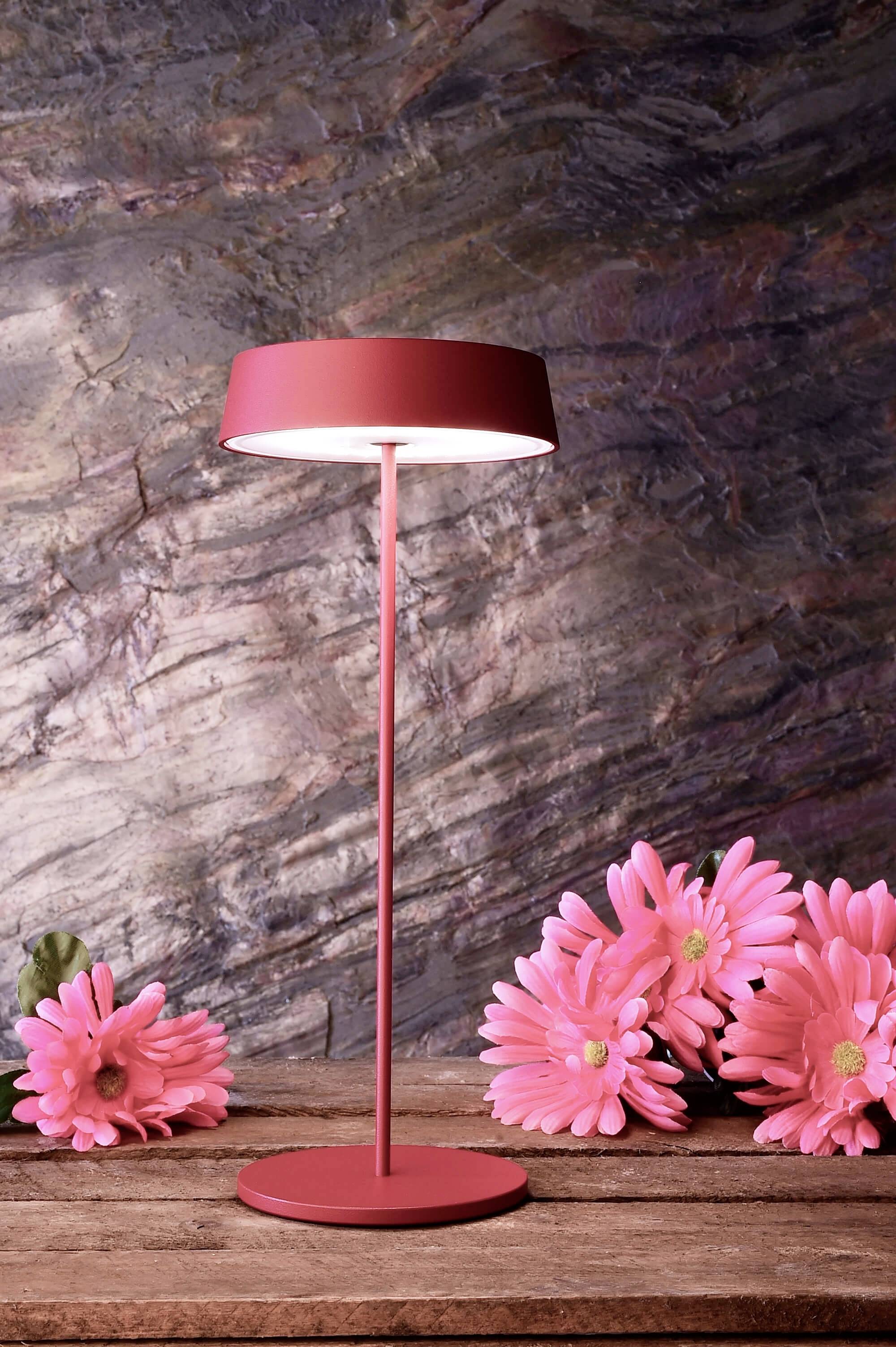 Rød bordlampe lyser ved siden af lyserøde blomster på et træbord foran en stenmur.