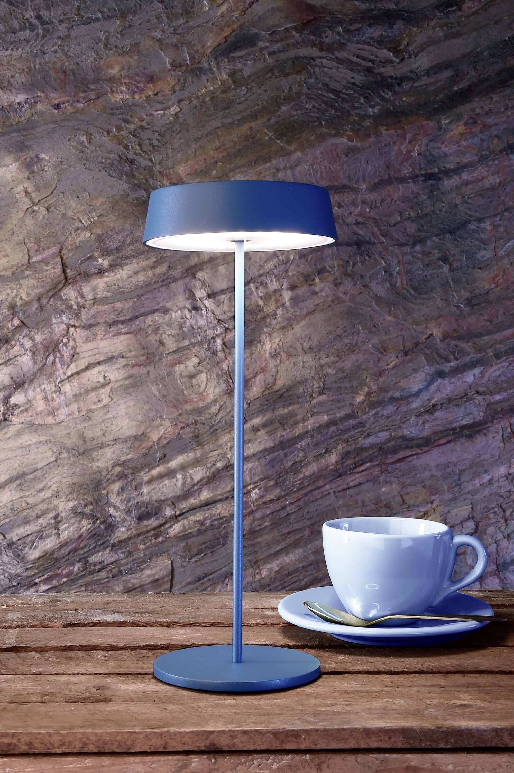 En blå bordlampe står tændt på et træbord ved siden af en blå kop med underkop foran en struktureret stenmur.