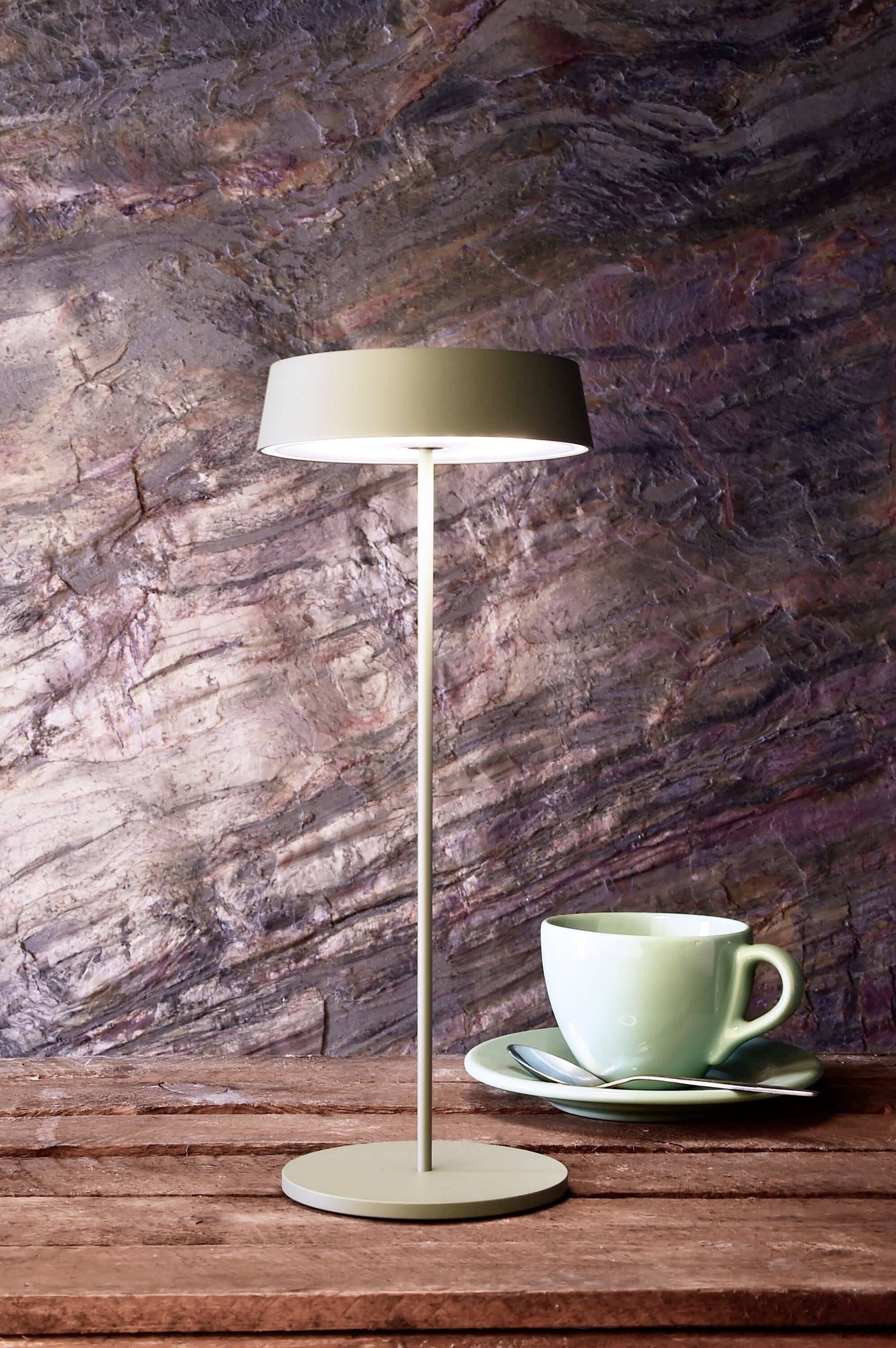 Et moderne bordlampe lyser op på et træbord. Ved siden af står en grøn kop med underkop foran en stenmur.