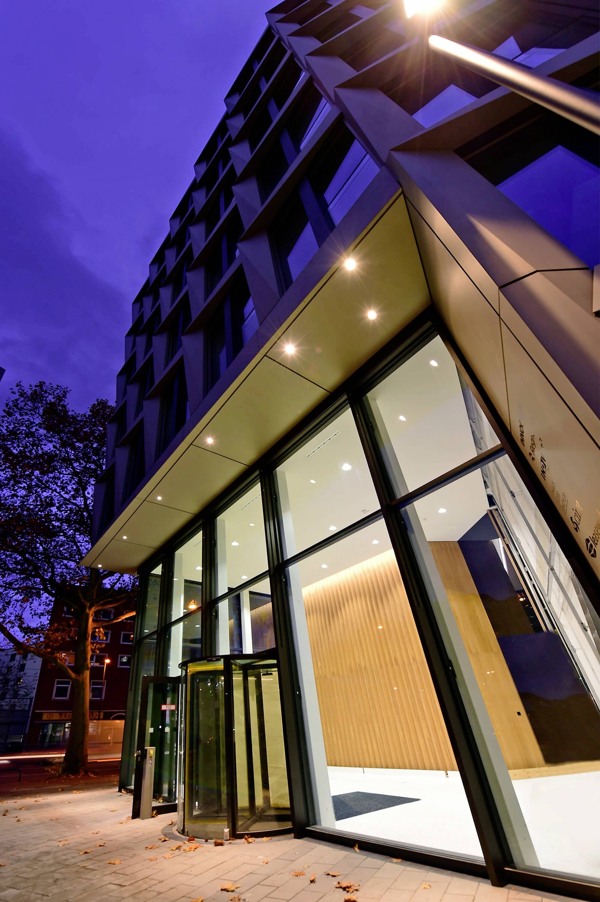 Moderne kontorbygnings indgang ved skumringen, med glasfacade og belysning. Et træ og boligbygninger i baggrunden.
