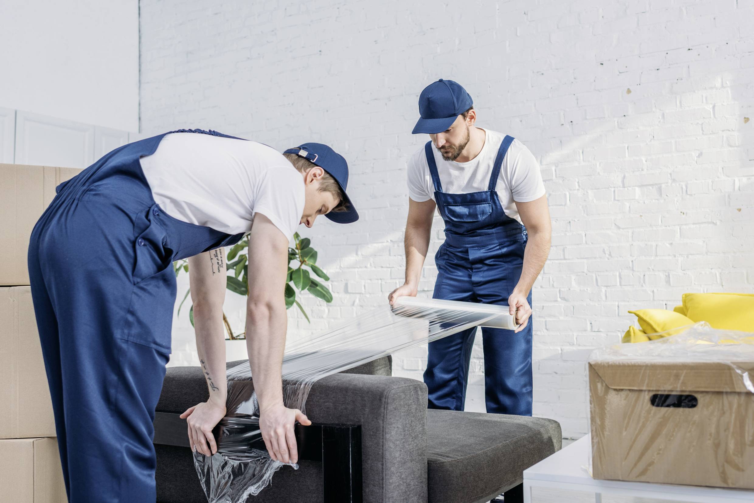 To mænd i arbejdstøj pakker en sofa ind i plastikfolie i et rum fyldt med pakkasser, tilsyneladende midt i en flytning.