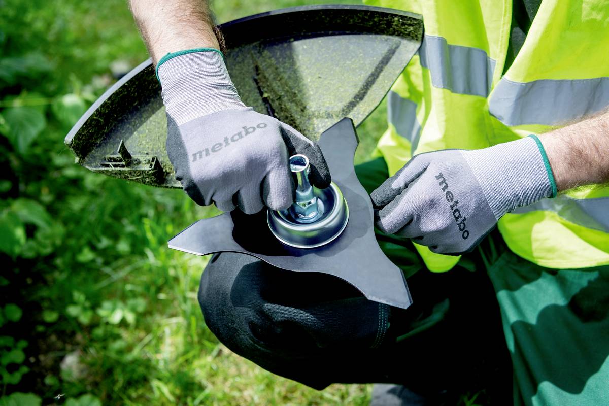 En person iført sikkerhedsvest og handsker reparerer eller udskifter et skæreredskab på en havemaskine udendørs.