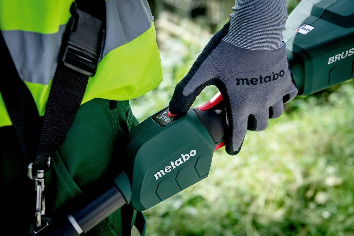 En person bærer handsker og holder et elektrisk haveredskab med påskriften 'metabo' i en grøn omgivelse.