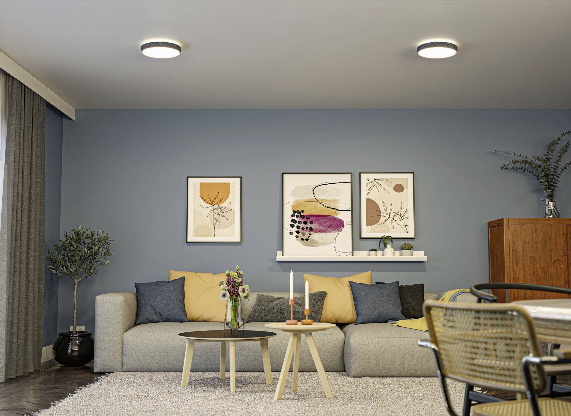 Helles Wohnzimmer med grå sofa, farvefulde puder, to runde borde med stearinlys og blomster, blå væg med kunstværker og planter.
