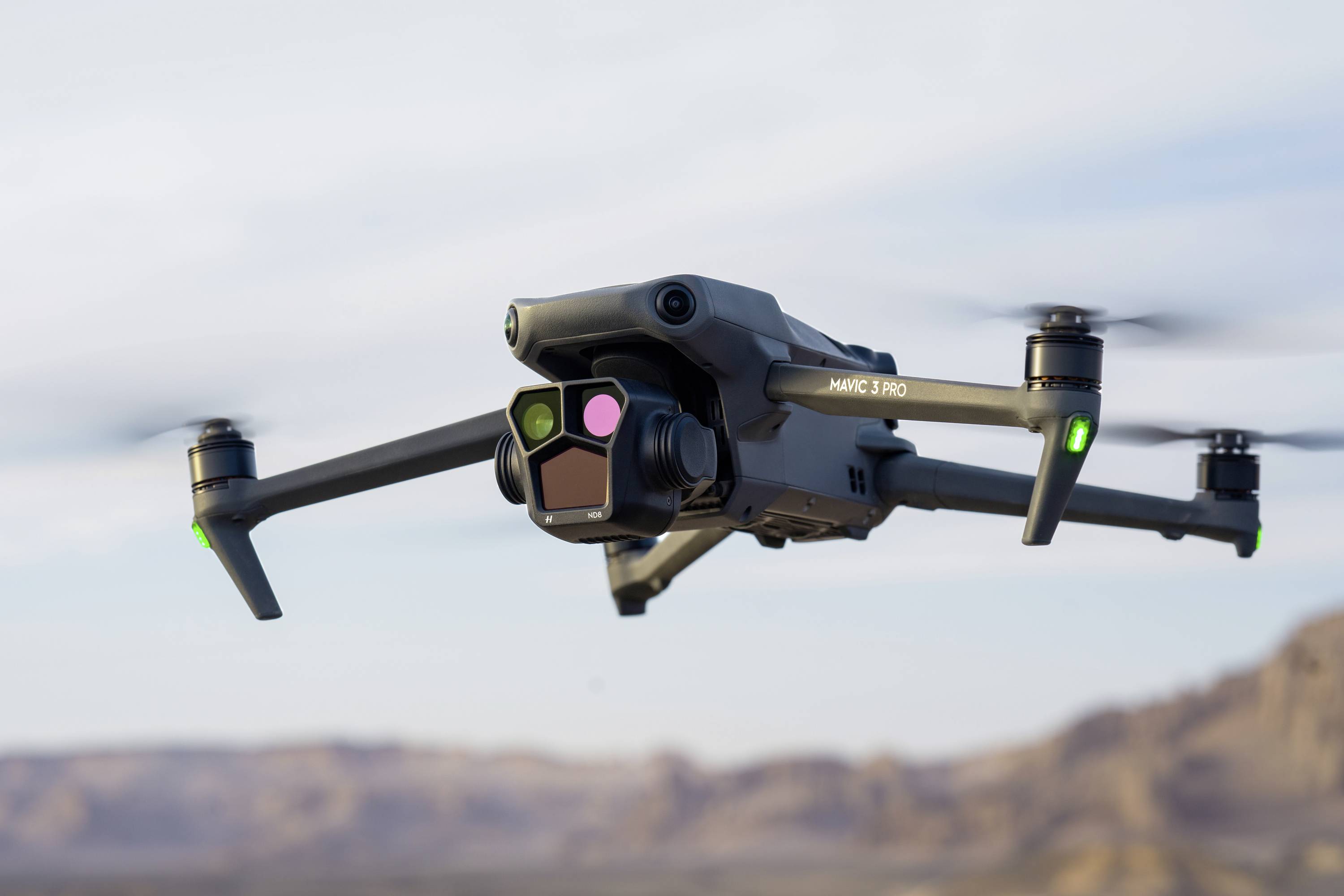 En drone svæver i luften over et øde ørkelandskab. Den har grønne positionslys og et kamera monteret fortil.