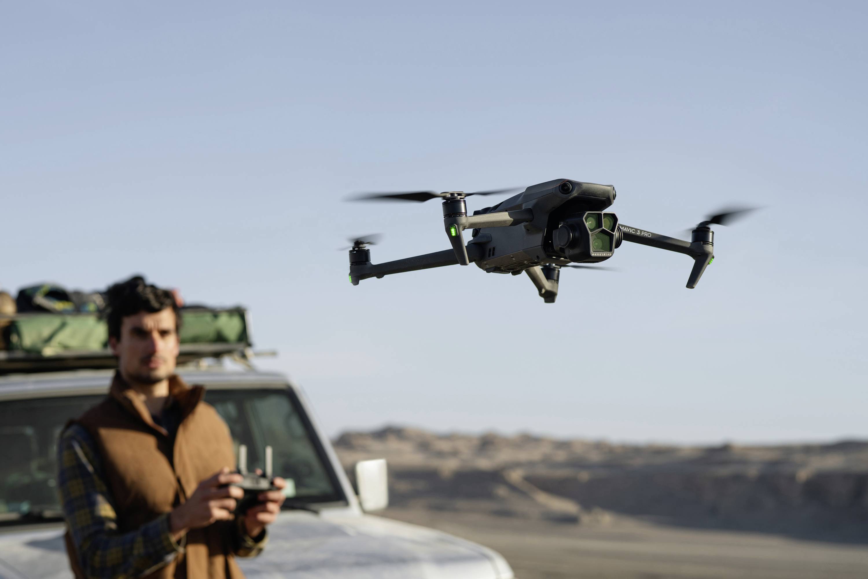 En mand styrer en svævende drone med en fjernbetjening i et øde ørkenet landskab. I baggrunden holder en terrængående bil.
