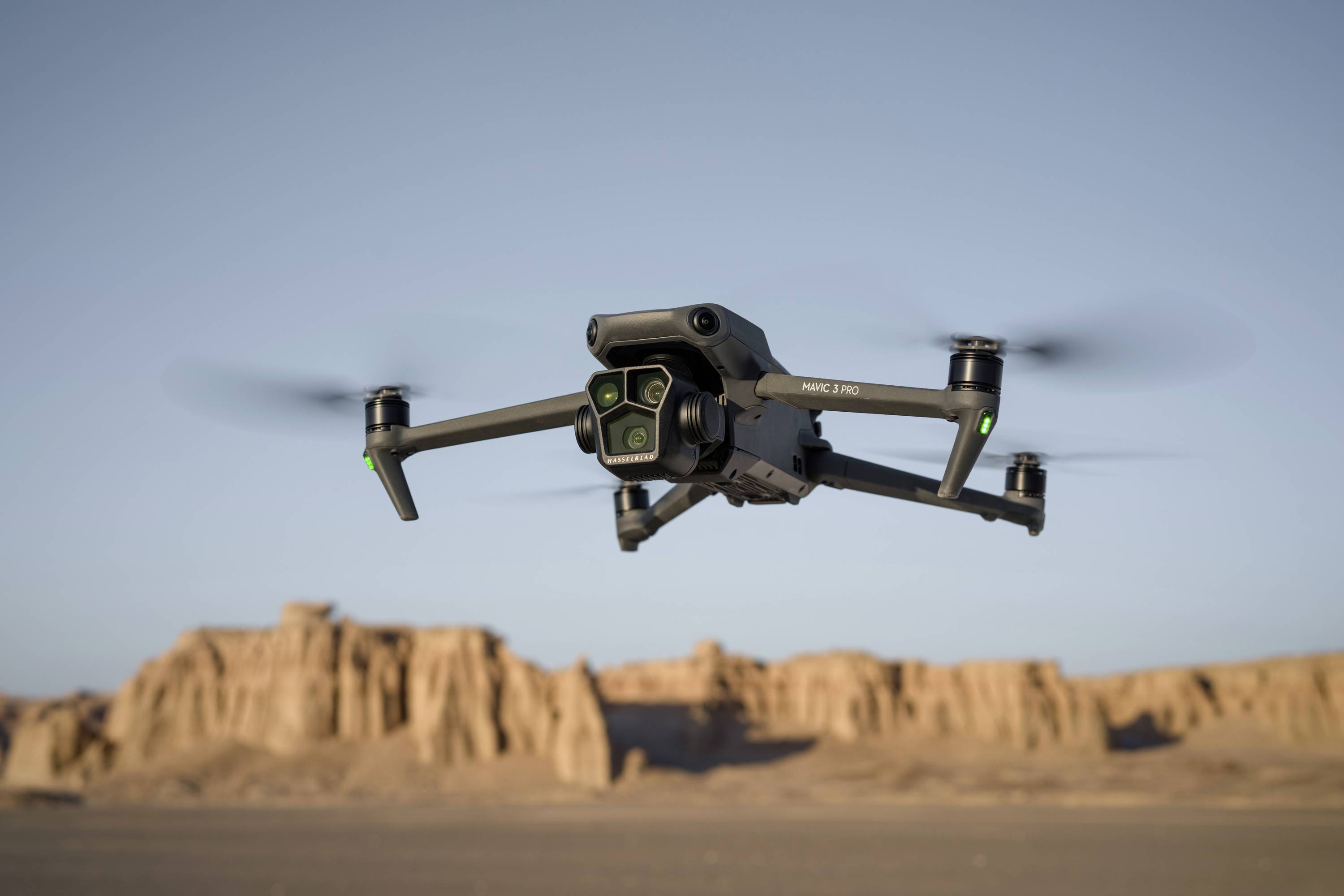En drone flyver i ørkenen, hvor klippeformationer ses i baggrunden under en klar himmel.