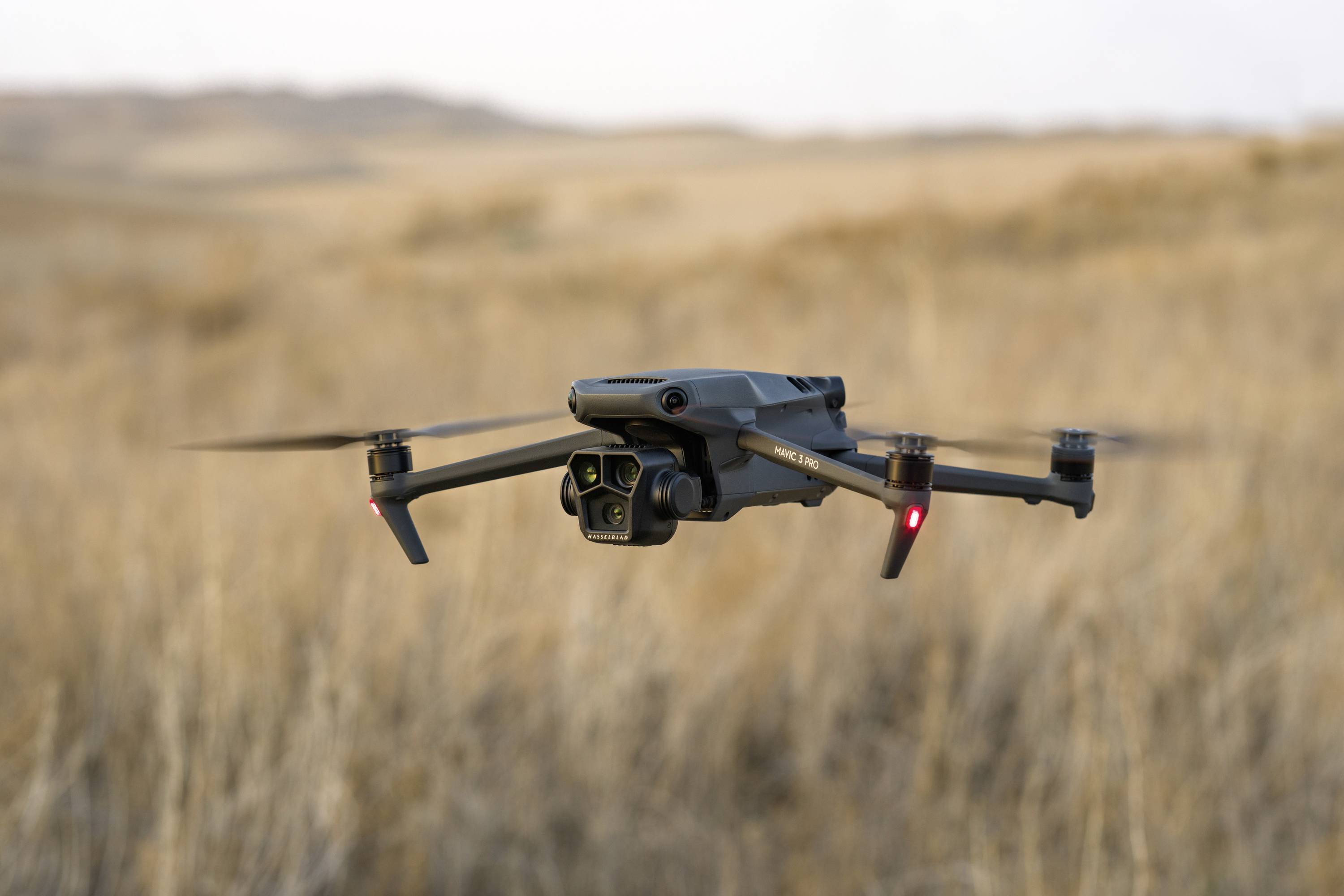 En drone svæver over en brun mark. Den har fire rotorer og et kamera. Baggrunden er et åbent landskab.