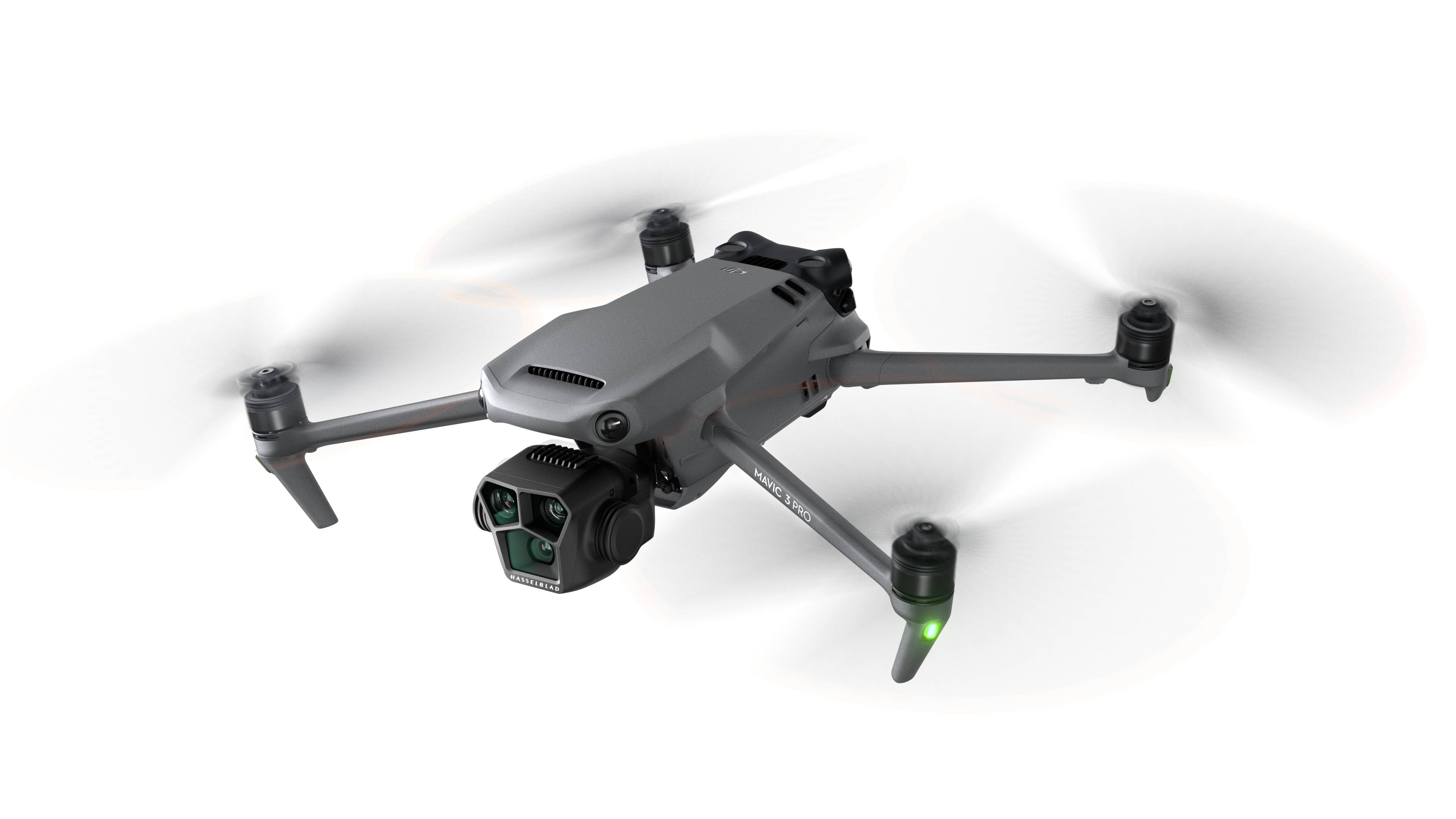 Grå drone i luften med fire rotorer og et kamera foran, propeller slørede ved bevægelse, hvid baggrund.
