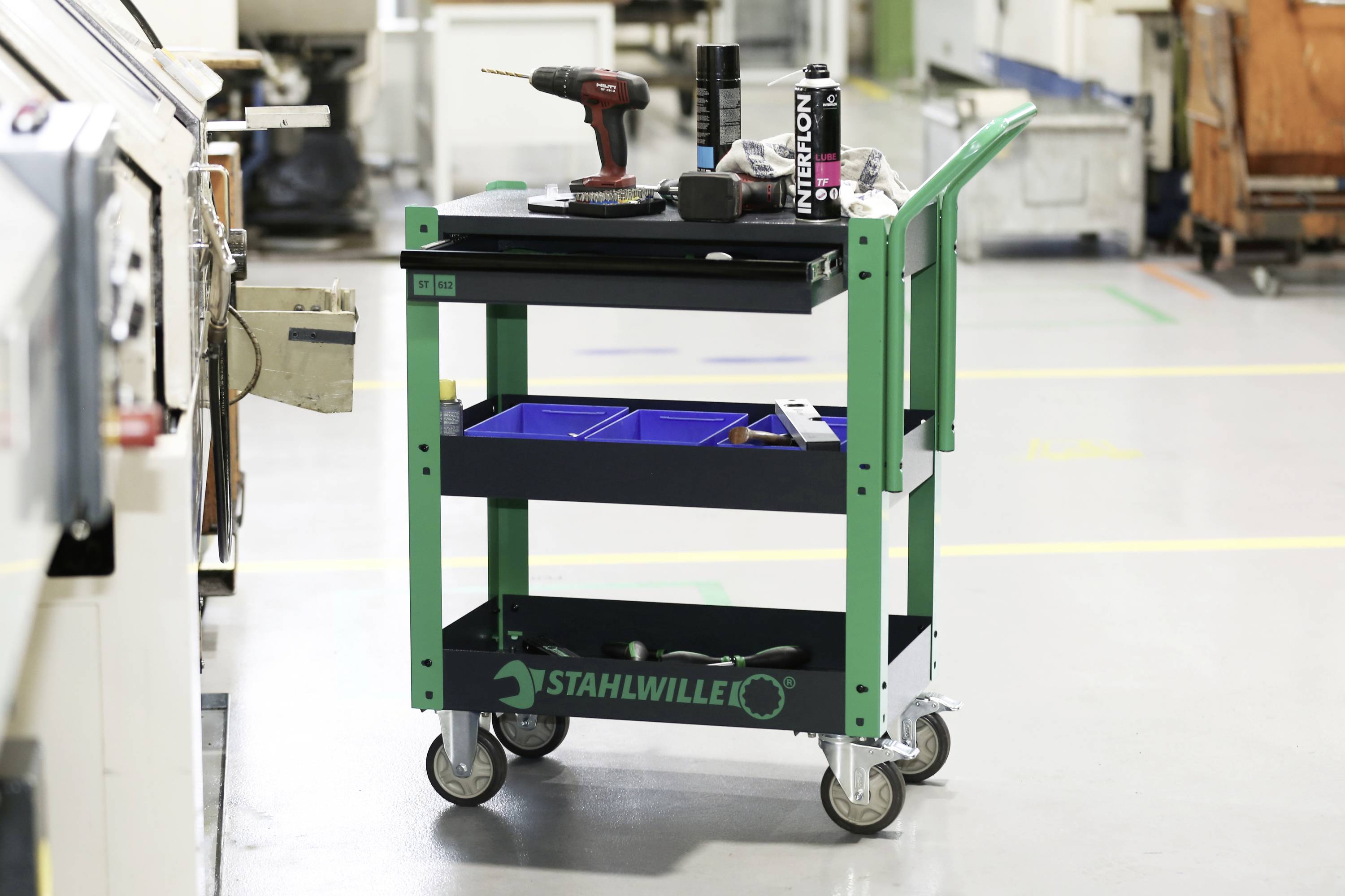 En grøn værktøjsvogn med tre hylder står i et værksted. På den øverste hylde ligger en skruetrækker og forskellige værktøjsdele.
