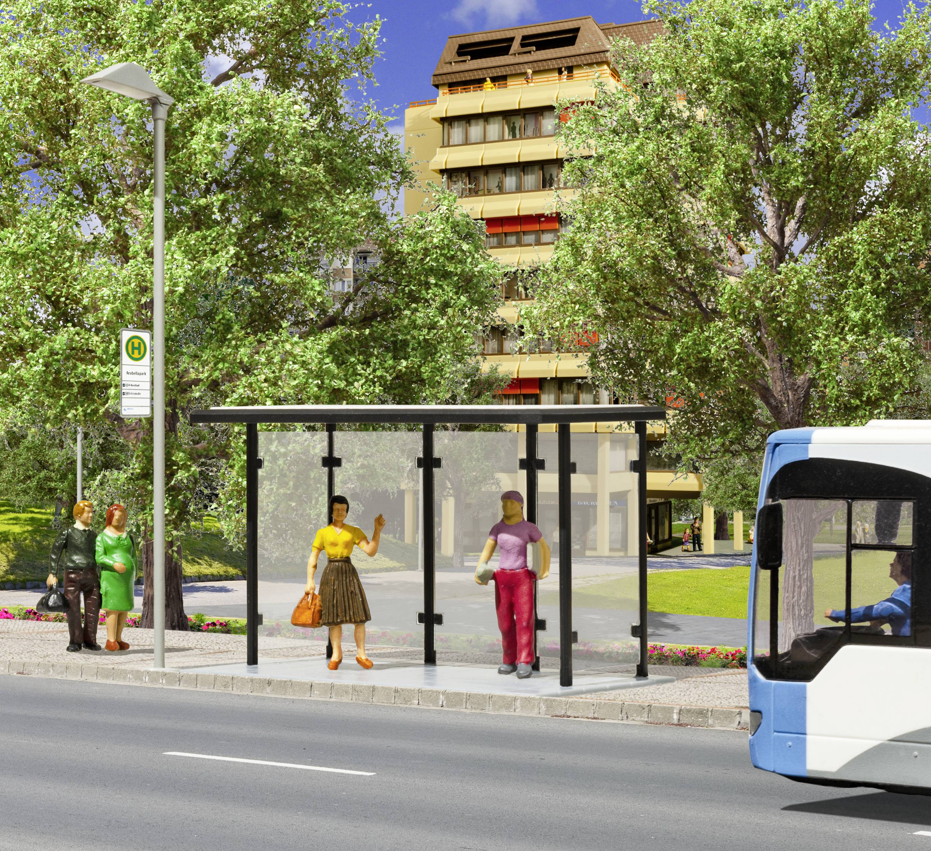 Busholdeplads med ventende personer, en forbikørende bus og en beboelsesbygning i baggrunden. Træer omkranser vejen.