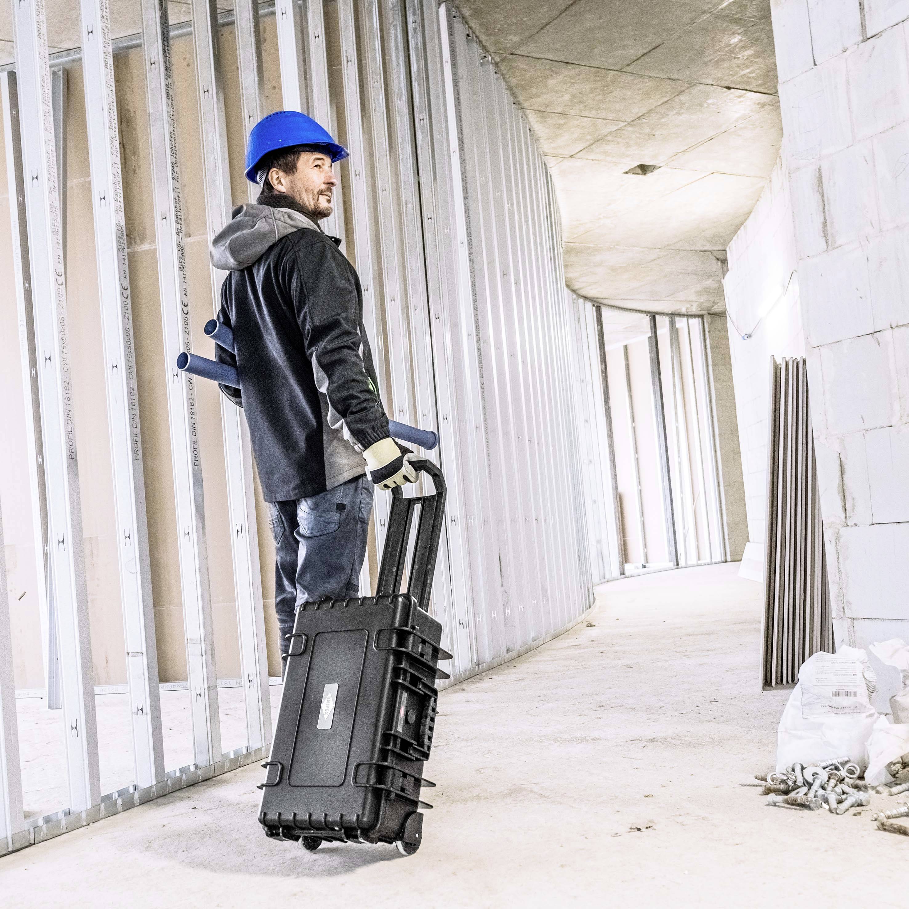 En bygningsarbejder med en blå sikkerhedshjelm trækker en sort værktøjskasse gennem en ufærdig bygningsgang med metalstænger.