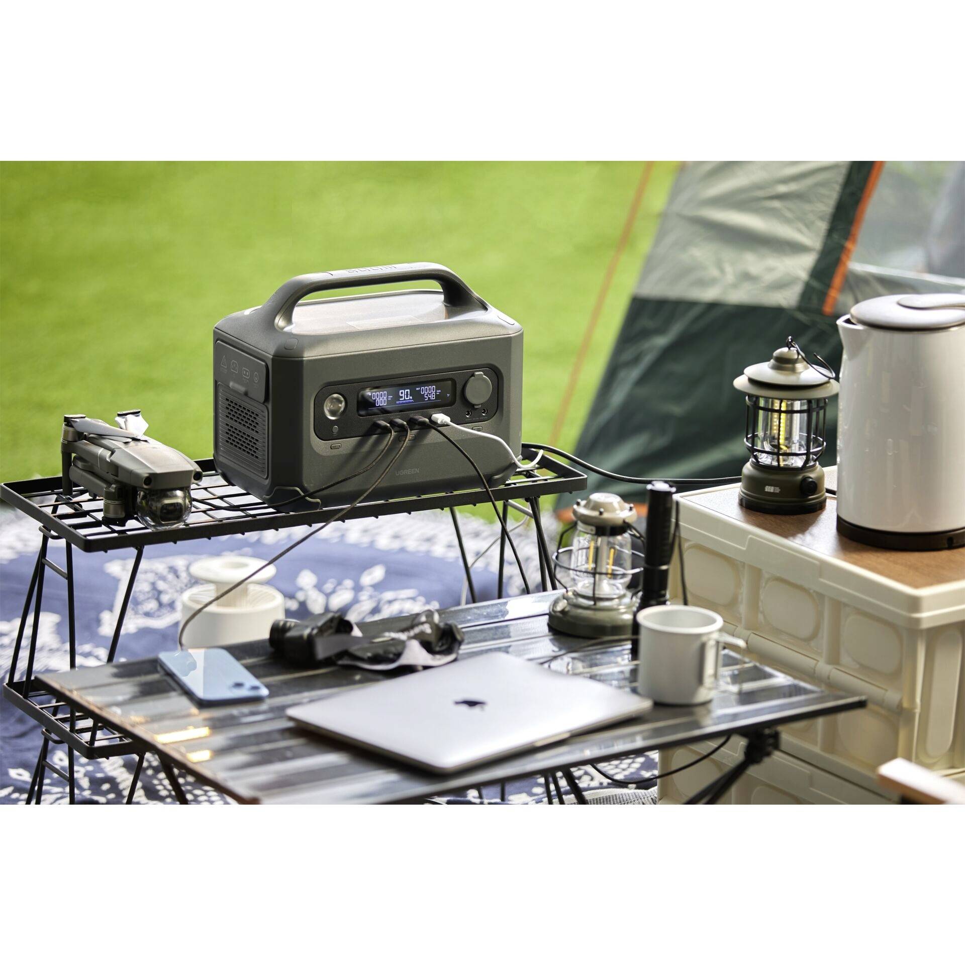 Campingscene med bærbar strømstation på et bord. Forskellige enheder som kamera, laptop og lampe er tilsluttet. I baggrunden står et telt.