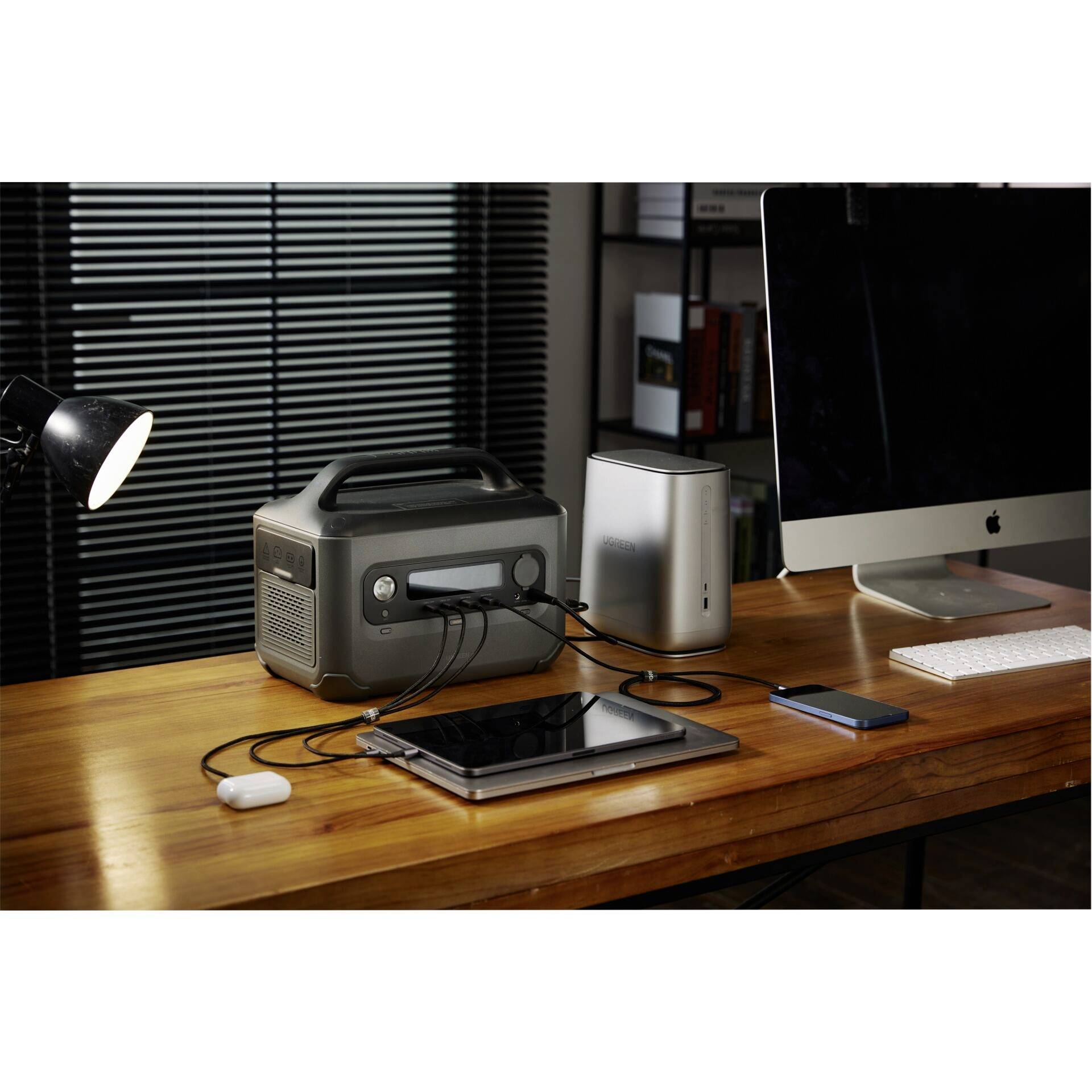 Et skrivebord med en computer, en lampe og flere elektroniske enheder, der er forbundet til en bærbar strømstation via kabler.