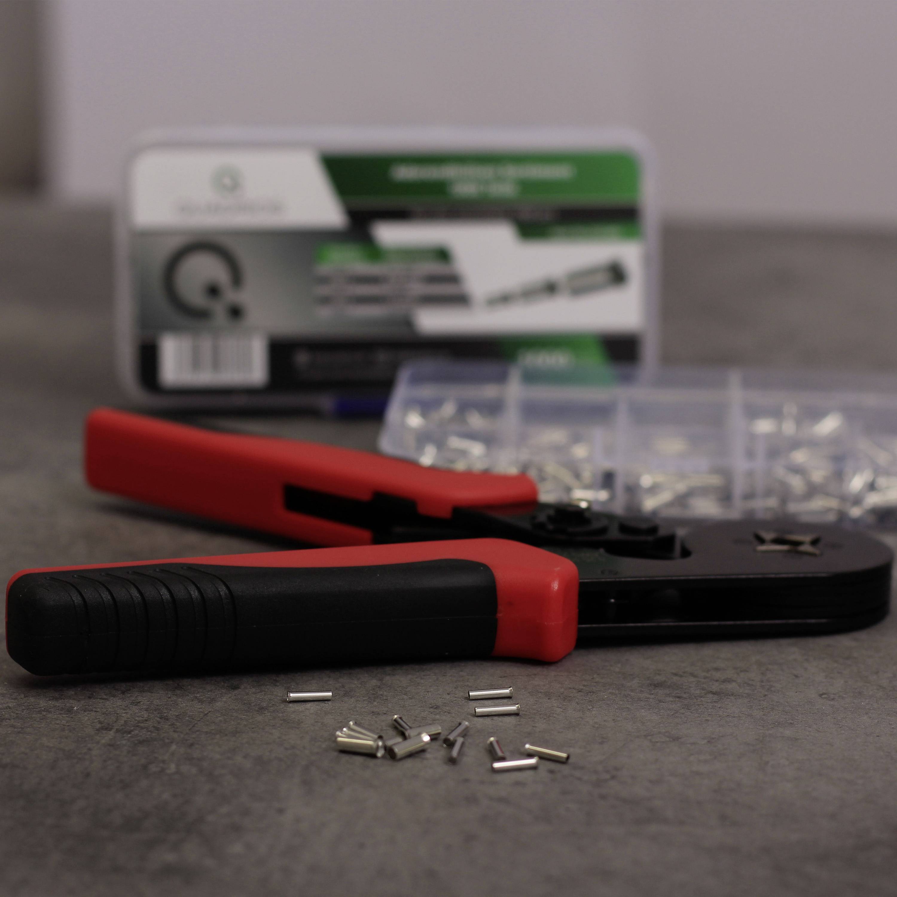 En rød crimptang ligger på et gråt bord ved siden af en åben beholder med metalclips. I baggrunden ses en mærket kasse.