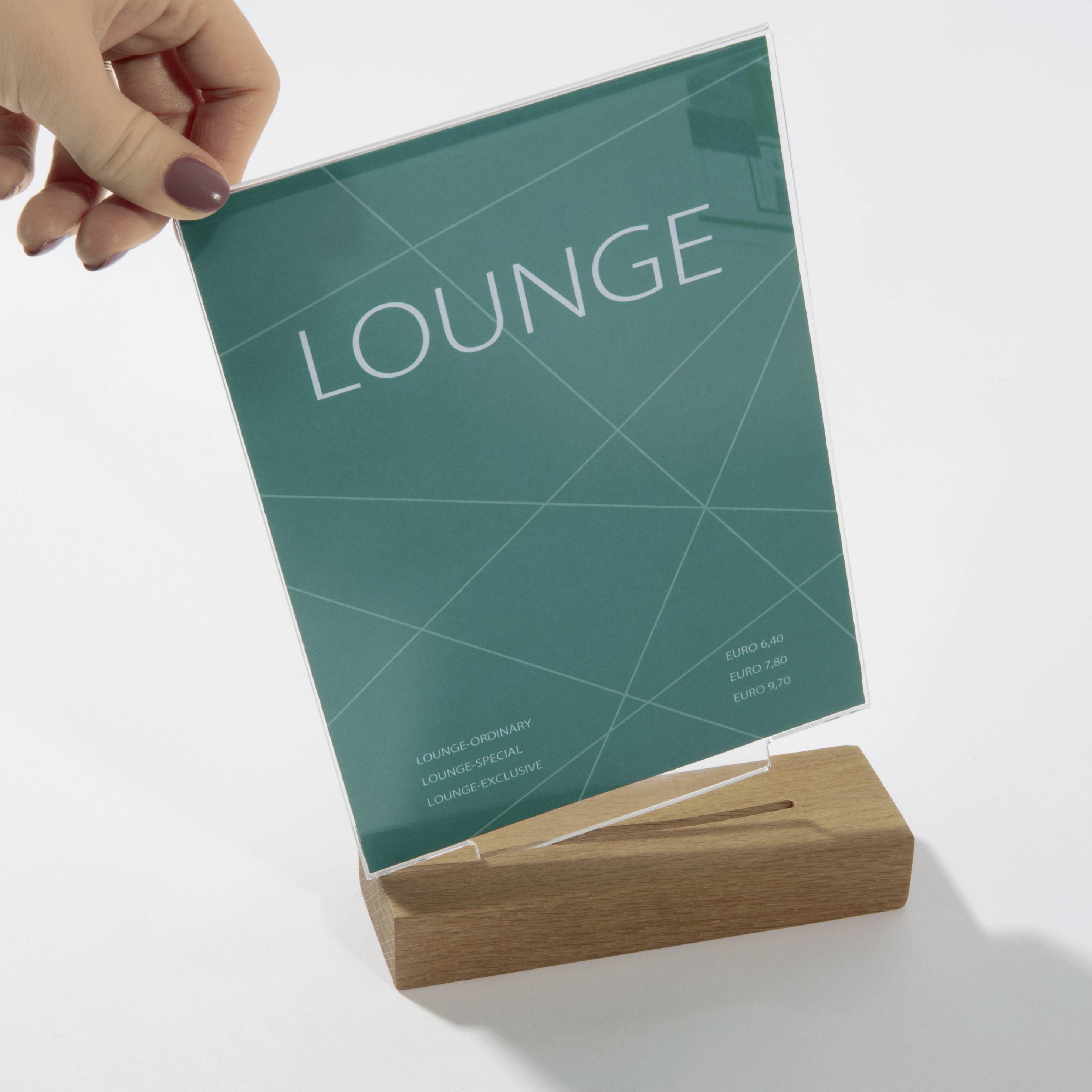 En hånd holder et grønt skilt med 'LOUNGE' og prisoplysninger. Det står i en træmonter.