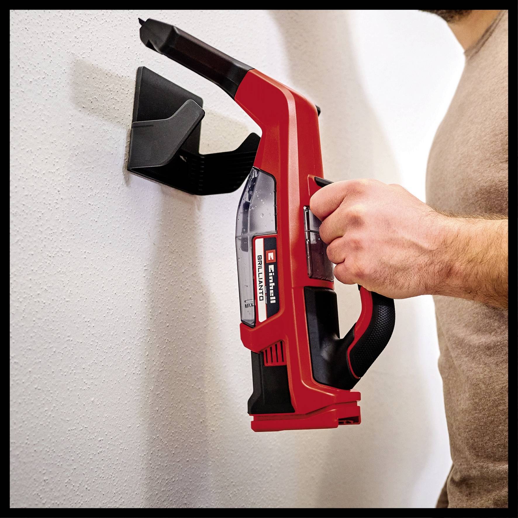 En person holder en rød, trådløs håndstøvsuger, der er monteret på en væg. Støvsugeren er fastgjort i en holder.