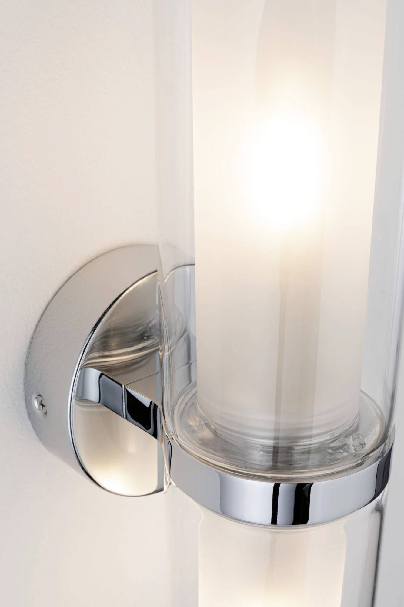 Væglampe med blank, forkromet holder og cylinderformet mælkehvidt glas, som udsender varmt lys.
