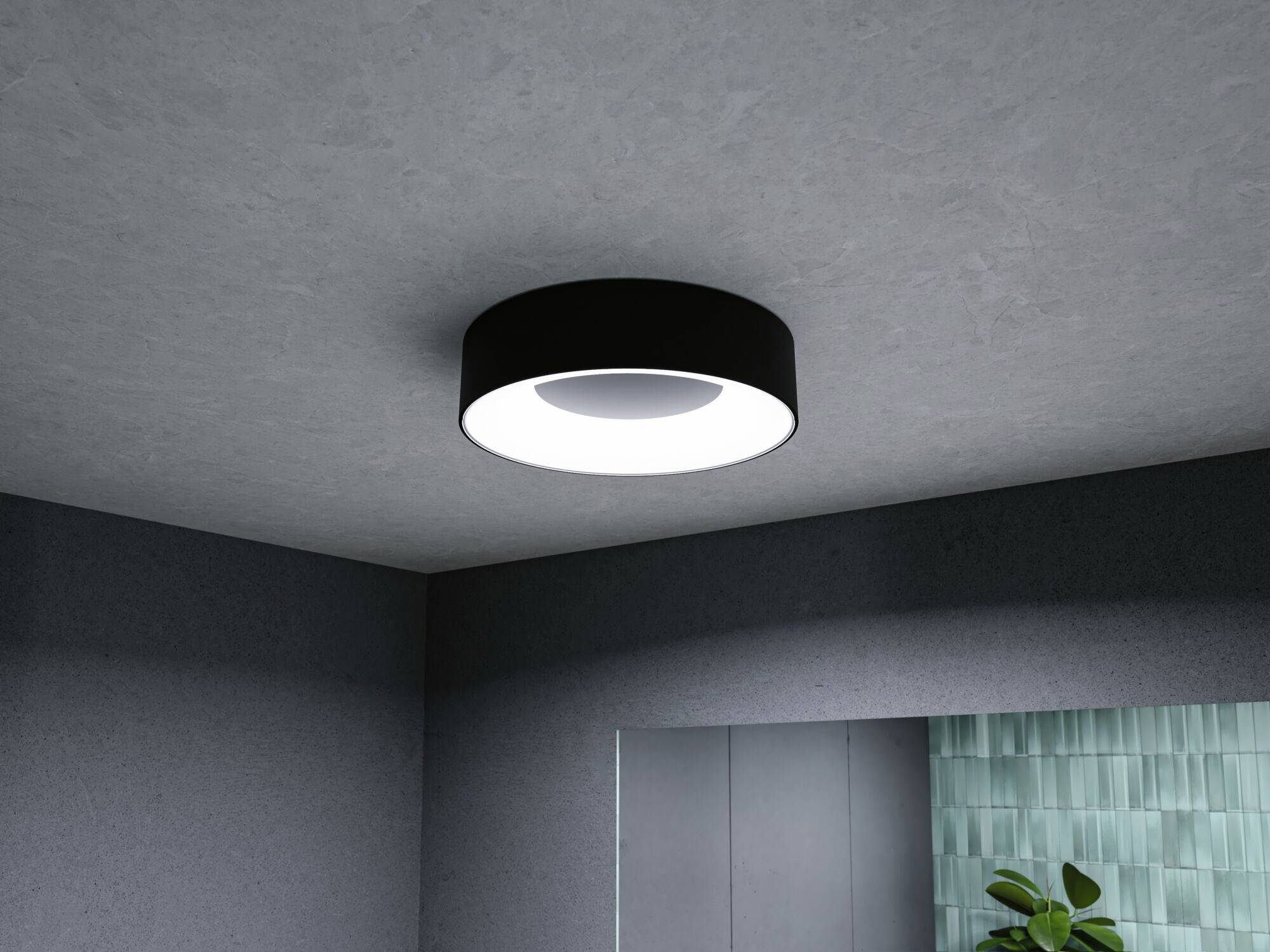 En rund loftslampe i moderne design lyser klart i et rum med grå vægge og et dekorativt grønt element.