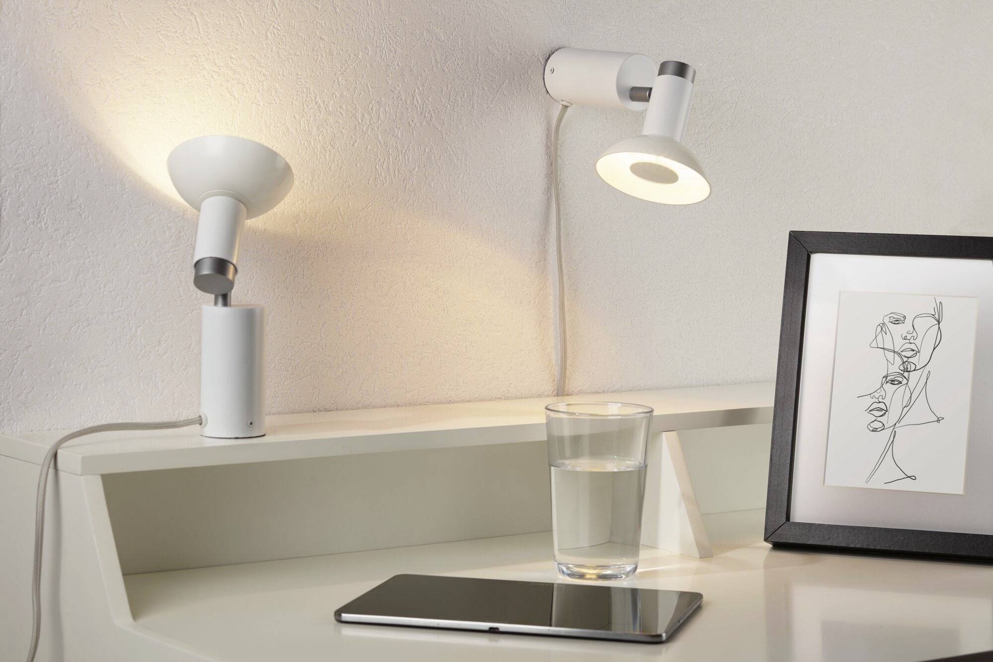 Skrivebord med to lamper, et vandglas, en smartphone og et indrammet kunstværk. Stemningen af en moderne, minimalistisk arbejdsflade.