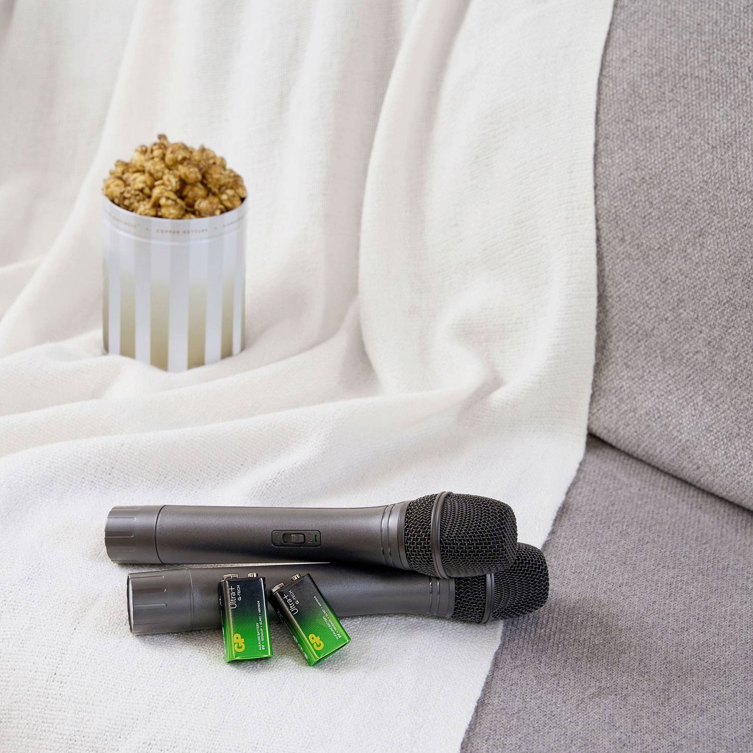 To kabelløse mikrofoner med batterier på et hvidt dække; ved siden af står en kop popcorn på en sofa.