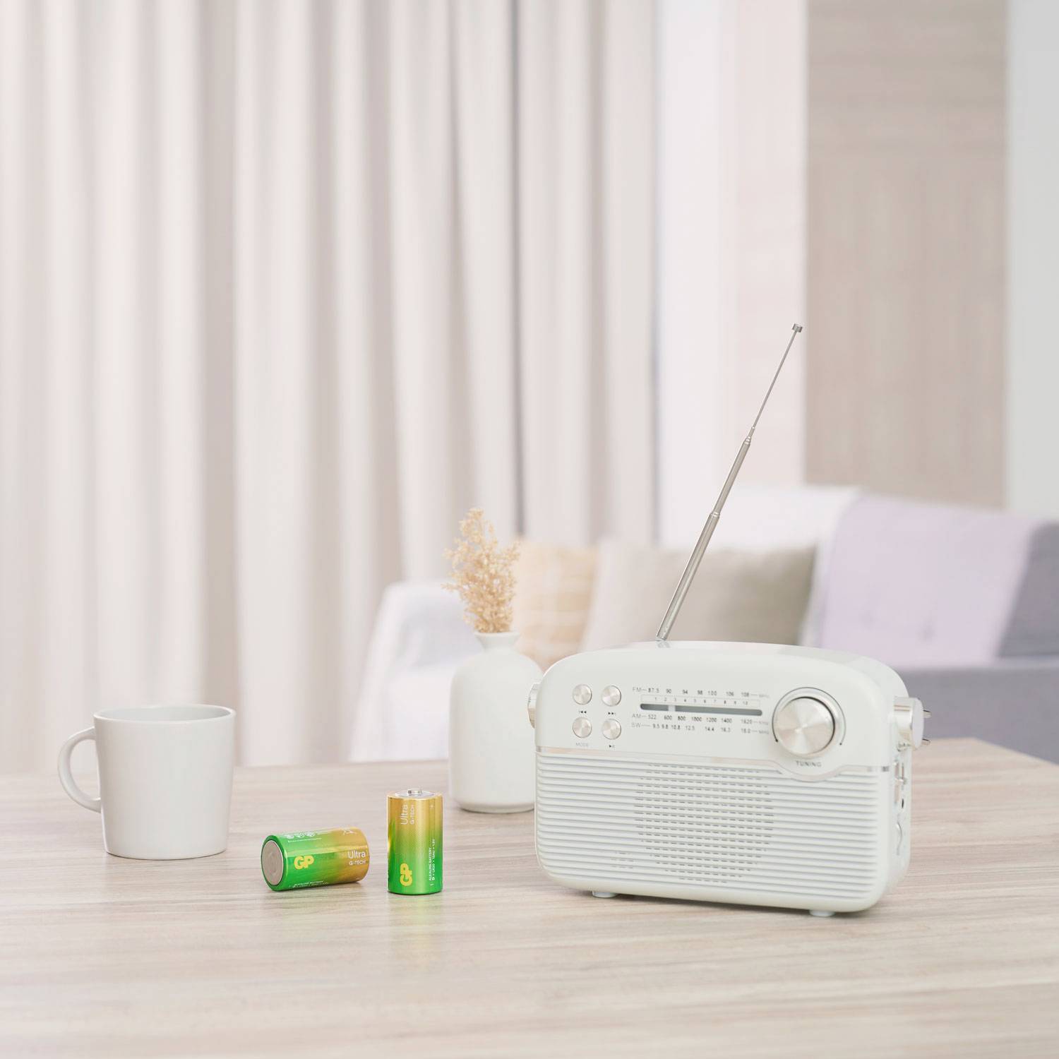 Hvidt retro-radio på et bord ved siden af en kop og to batterier i et lyst stueområde.