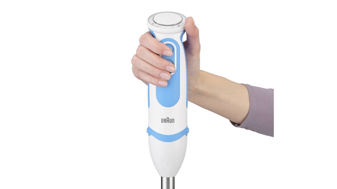 En person holder en hvid stavblender med et blåt håndtag. Blendermixeren har et moderne design, og mærket er synligt.