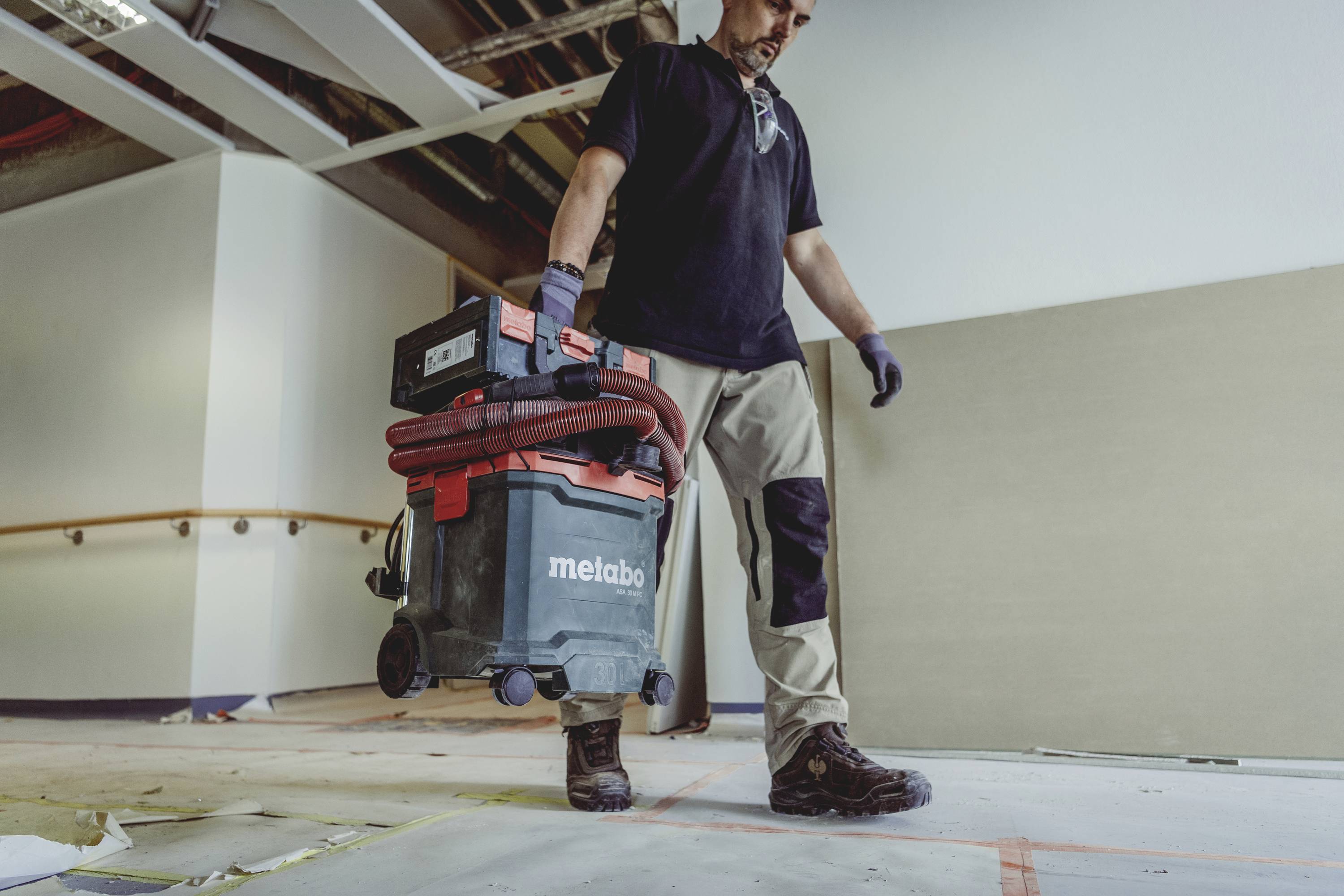 En arbejder bærer en industriel støvsuger hen over et gulv på en byggeplads. I baggrunden er blottede loftsstrukturer synlige.
