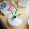 To blomlignende Lego-blomster i en hvid vase på et træbord, dekorativt arrangeret foran en uskarp baggrund.