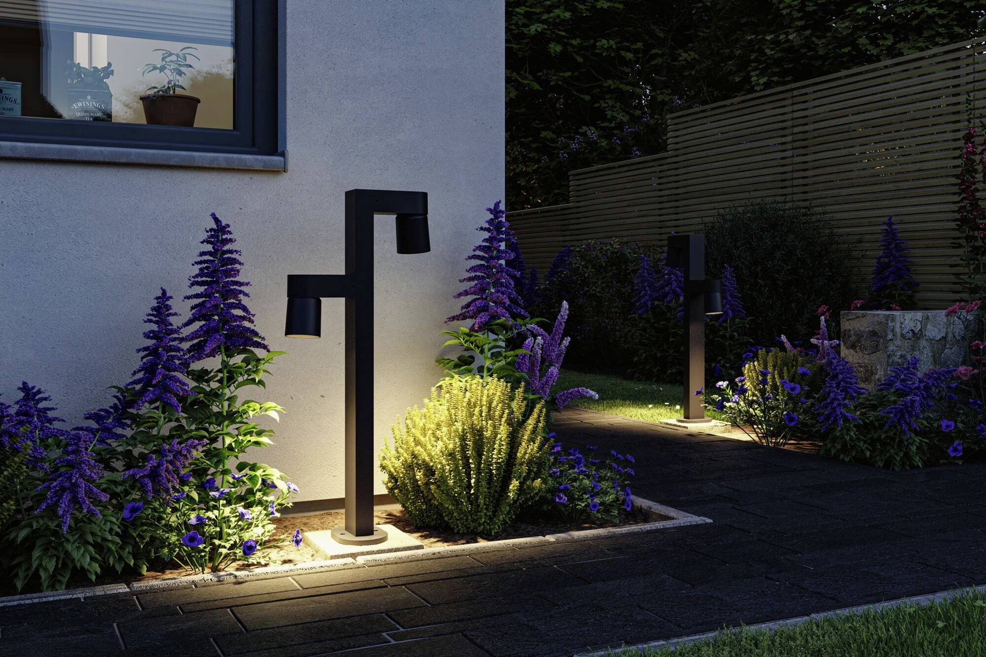 En moderne havbelysning oplyser stien langs et velplejet blomsterbed med lilla planter i en natlig have.