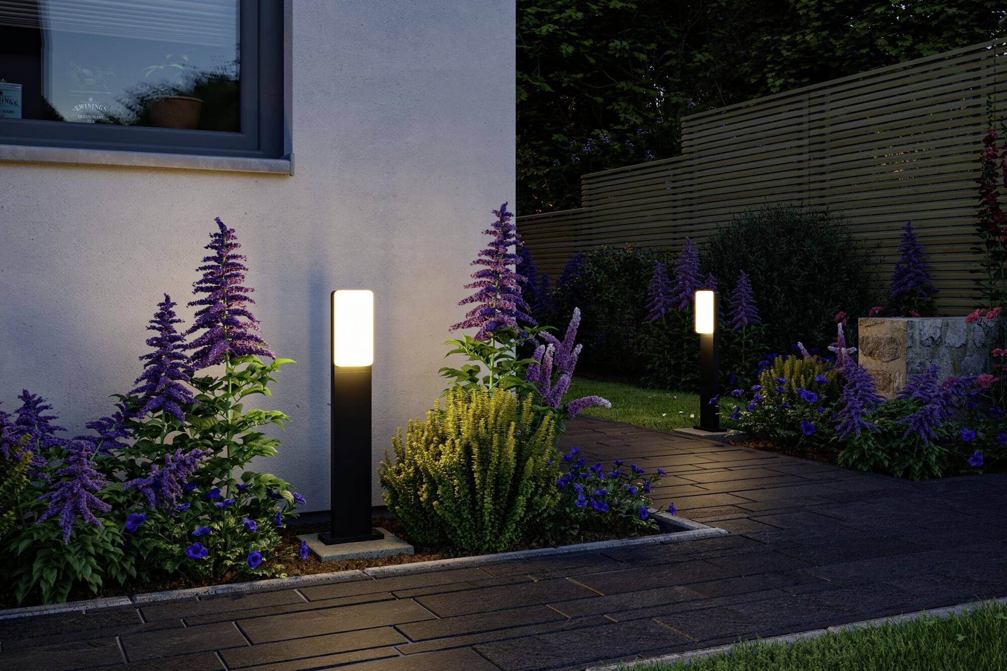 To belyste havelygter belyser en sti, der er kantet med lilla blomster. I baggrunden ses et træhegn.