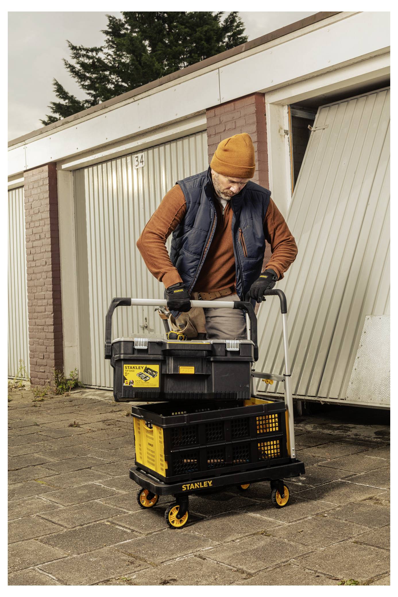 En person i det fri, der bærer en orange strikket hat og handsker, organiserer forskellige værktøjer i sorte og gule værktøjskasser på en rullevogn.