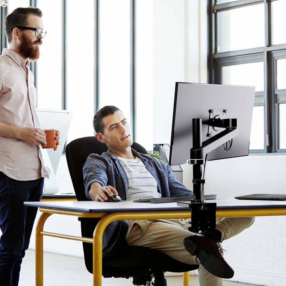 To mænd på kontoret, hvor den ene sidder ved skrivebordet foran en computer, mens den anden står og holder en kop. Lys, moderne omgivelser.